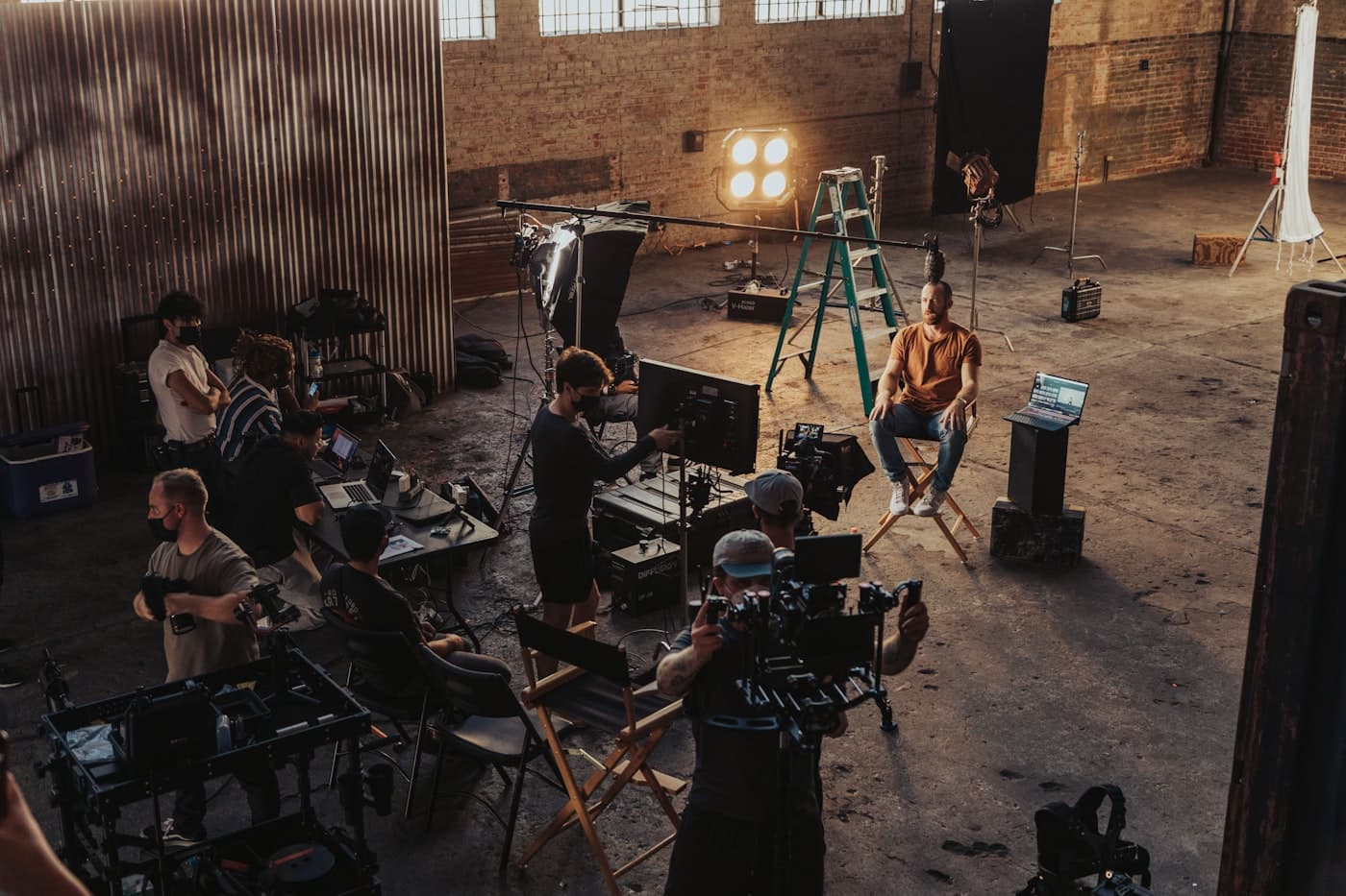 A film crew setting up lighting and camera equipment around a seated subject in a warehouse studio